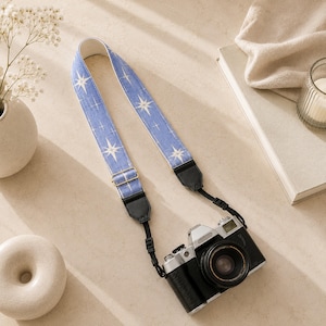 May include: A camera strap featuring a periwinkle blue fabric with a repeating white starburst pattern. The strap includes gold-toned metal hardware for length adjustment and black leather-like end tabs that attach to a silver and black camera body.