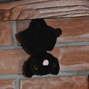 Può includere: Un pipistrello di peluche lavorato all'uncinetto di colore nero appeso a testa in giù. Il pipistrello presenta una consistenza morbida e soffice, due grandi occhi color ambra e un piccolo naso rosa chiaro. Le ali sono ripiegate contro il corpo.