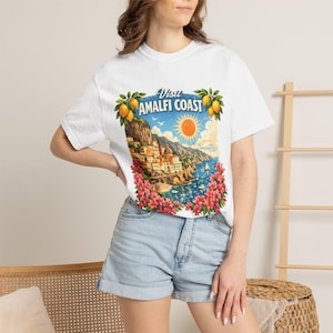 Może przedstawiać: Biały t-shirt z krótkim rękawem z kolorowym nadrukiem graficznym przedstawiającym Wybrzeże Amalfi. Wzór zawiera nadmorskie miasteczko, jasnopomarańczowe słońce, błękitną wodę i różowe kwiaty, z nadrukowanym u góry napisem "Visit AMALFI COAST".