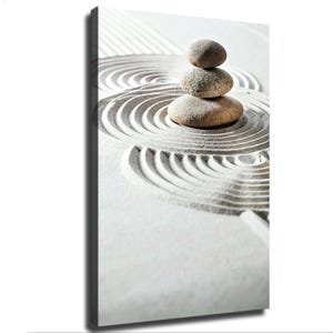 May include: A vertical canvas print featuring a stack of three smooth, light brown stones balanced on white sand. The sand is raked into concentric circular patterns that radiate outward from the base of the stone stack, creating a textured effect.