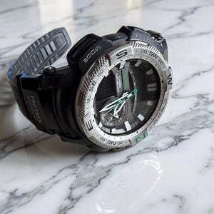 May include: A black Casio Pro Trek watch with a silver-toned rotating bezel featuring compass directions. The watch face displays white hour markers, green-accented hands, and the text Casio Twin Sensor. It has a black resin strap.