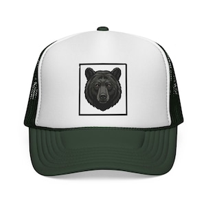 Può includere: Un cappellino trucker con pannello frontale bianco e una grafica quadrata nera contenente un'illustrazione dettagliata di una testa di orso nero. Il cappellino presenta una visiera verde scuro e una parte posteriore in rete nera strutturata.