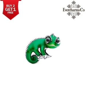May include: A silver-toned charm shaped like a chameleon with vibrant green enamel detailing, featuring large black eyes and a curled tail. The charm has a textured, beaded spine and a smooth finish on the body, presented against a plain background.