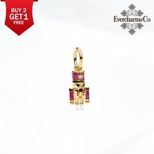 May include: A gold-toned nutcracker charm featuring pink glitter enamel details on the hat and sleeves, a small clear stone on the hat, and white enamel accents on the feet. The charm includes a circular jump ring for attachment to jewelry.