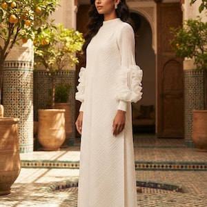 Könnte beinhalten: Ein bodenlanges weißes Kleid mit hohem Ausschnitt, langen Ärmeln mit voluminösen Rüschenbündchen und einer dezenten diagonalen Plisseestruktur im gesamten Stoff. Das Kleidungsstück hat eine gerade Silhouette und reicht bis zum Boden.
