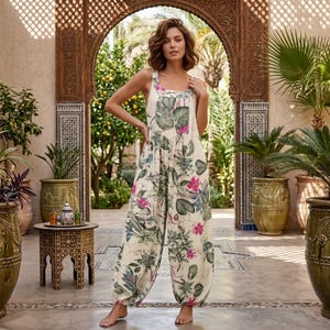 May include: A sleeveless, loose-fitting jumpsuit featuring a cream-coloured base with a vibrant botanical print of large green tropical leaves and bright pink flowers. The garment has a scoop neckline and tapered, gathered ankles at the hem.