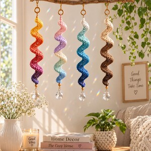 Op de afbeelding: Vier spiraalvormige gehaakte ornamenten hangen aan een houten tak. Elk heeft een kleurverloop, waaronder geel-paars, roze-blauw, wit-donkerblauw en crème-bruin, afgewerkt met een heldere gefacetteerde kristallen kraal aan de onderkant.