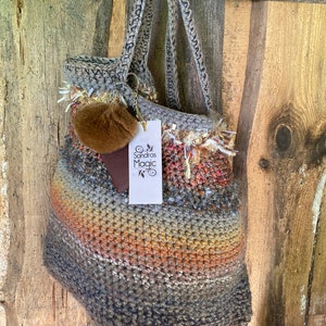 May include: A crocheted tote bag with a brown and gray color scheme. The bag has a fluffy brown pom-pom attached to the handle. The bag is hanging on a wooden wall with a tag that says "Sandra's Magic".