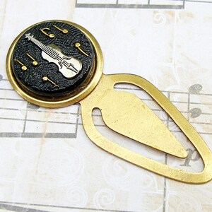 May include: A gold bookmark with a black and white violin and musical notes on a round, black button. The bookmark is on a sheet of music.