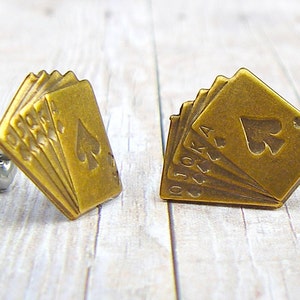 May include: A pair of gold-toned earrings featuring a design of playing cards with the ace of spades on top. The cards are fanned out and the earrings have a stud back.