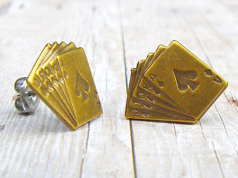 May include: A pair of gold-toned earrings featuring a design of playing cards with the ace of spades on top. The cards are fanned out and the earrings have a stud back.