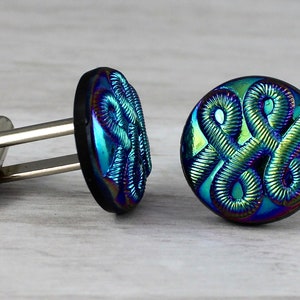 Op de afbeelding: Een paar manchetknopen met een zwarte basis en iriserende blauwe en groene glazen toppen. De glazen toppen hebben een reliëf, ingewikkeld ontwerp van verweven lijnen.
