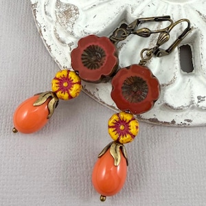 Może przedstawiać: Ręcznie robione kolczyki z kwiatowym wzorem. Kolczyki mają czerwone wierzchołki w kształcie maku, żółte i czerwone koraliki w kształcie kwiatów oraz pomarańczowe koraliki w kształcie łezki. Metalowe elementy mają wykończenie ze starego mosiądzu.