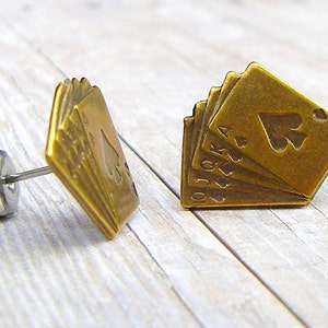 May include: A pair of gold-toned earrings featuring a design of playing cards, including the 10, Jack, Queen, King, and Ace of Spades.