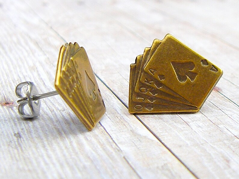 May include: A pair of gold-toned earrings featuring a design of playing cards, including the 10, Jack, Queen, King, and Ace of Spades.