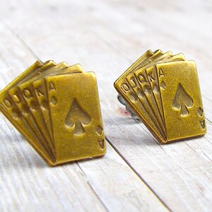 May include: A pair of gold-tone earrings featuring a design of five playing cards, including the Ace of Spades, 10, Jack, Queen, and King. The cards are arranged in a fan shape.