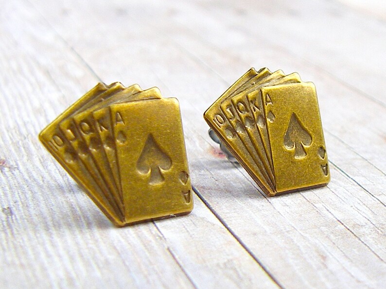 May include: A pair of gold-tone earrings featuring a design of five playing cards, including the Ace of Spades, 10, Jack, Queen, and King. The cards are arranged in a fan shape.
