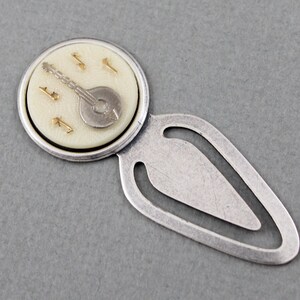 May include: Silver bookmark with a round, white, and silver charm featuring a banjo and musical notes. The bookmark is shaped like a teardrop with a loop at the top.