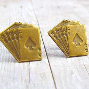 May include: A pair of gold-toned metal earrings shaped like a fan of playing cards. The cards are all spades and include the 10, Jack, Queen, King, and Ace.