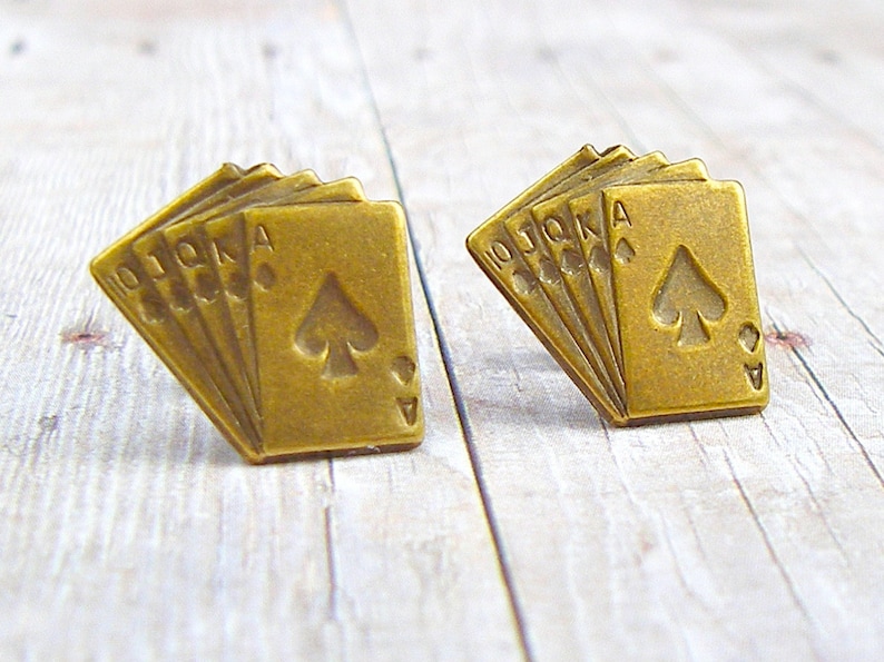 May include: A pair of gold-toned metal earrings shaped like a fan of playing cards. The cards are all spades and include the 10, Jack, Queen, King, and Ace.