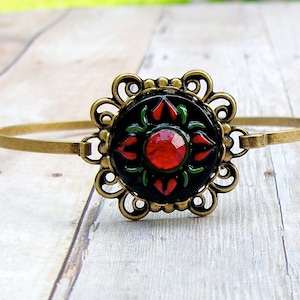 May include: A gold bangle bracelet with a black and green enamel floral design and a red rhinestone in the center.