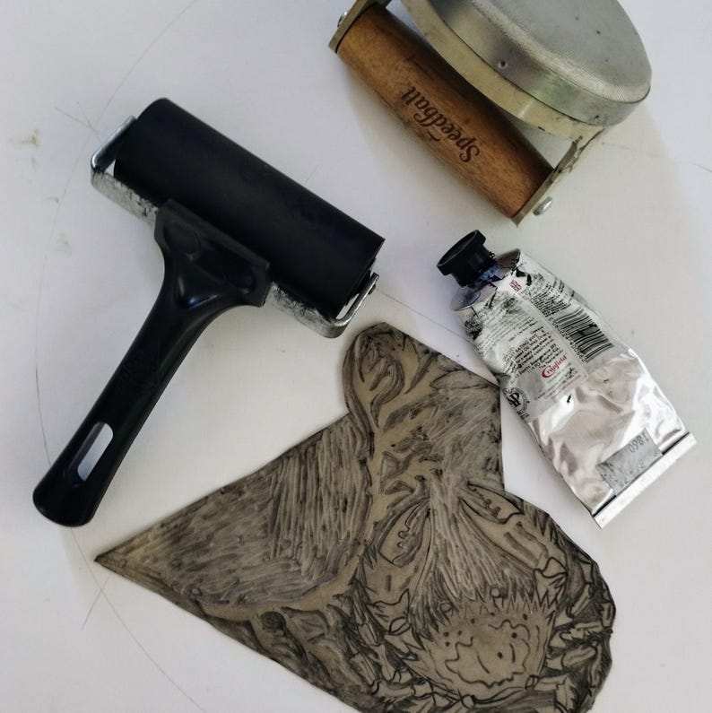 May include: A black rubber brayer, a wooden-handled Speedball brayer with a metal frame, a tube of Cranfield ink, and a carved linoleum block with an intricate design. These printmaking tools are arranged together for relief printing projects.