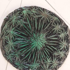 Pu&ograve; includere: Un piatto decorativo circolare caratterizzato da un motivo a raggiera in nero scuro e verde brillante. Il design &egrave; composto da numerose forme radianti e appuntite simili a foglie che si estendono dal centro verso il bordo esterno in modo denso.