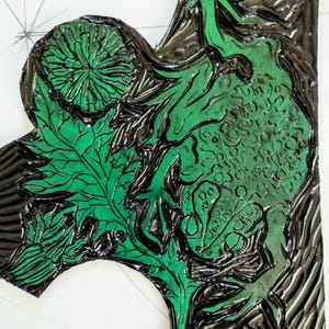 Pu&ograve; includere: Un blocco per linoleografia con inchiostro verde brillante applicato su una superficie nera intagliata. Il design presenta intricati motivi botanici, tra cui un fiore circolare, venature delle foglie dettagliate e forme organiche.
