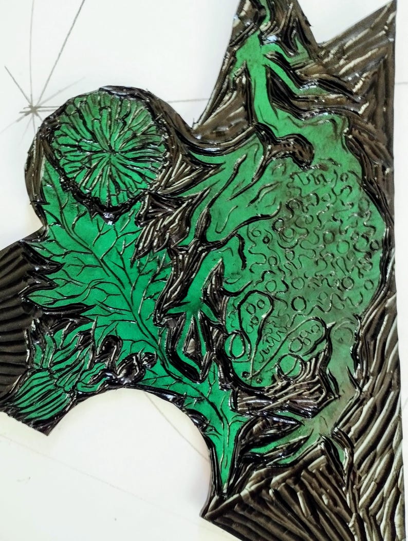 Pu&ograve; includere: Un blocco per linoleografia con inchiostro verde brillante applicato su una superficie nera intagliata. Il design presenta intricati motivi botanici, tra cui un fiore circolare, venature delle foglie dettagliate e forme organiche.