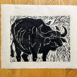 May include: A black and white linocut print on off-white paper featuring a buffalo with two small birds perched on its back. The artwork includes the text "Buffalo" at the bottom center, the edition number "1/20" on the left, and a signature on the right.