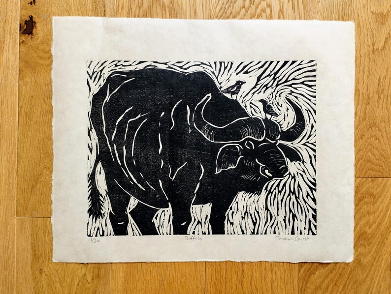 May include: A black and white linocut print on off-white paper featuring a buffalo with two small birds perched on its back. The artwork includes the text "Buffalo" at the bottom center, the edition number "1/20" on the left, and a signature on the right.