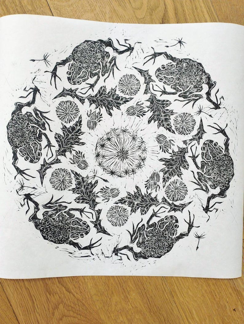 Circular black and white linocut art repeat print featuring textured toads arranged in a radial mandala design surrounded by dandelion flowers, leaves, and floating seeds on white Japanese washi paper.