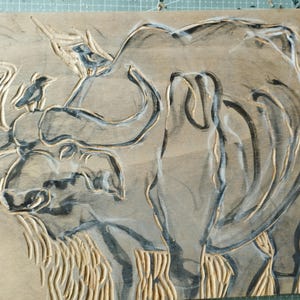 May include: A rectangular wooden block featuring a carved relief of a large buffalo with a small bird perched on its head. The design is highlighted with dark ink within the carved grooves, contrasting against the natural light brown wood surface.