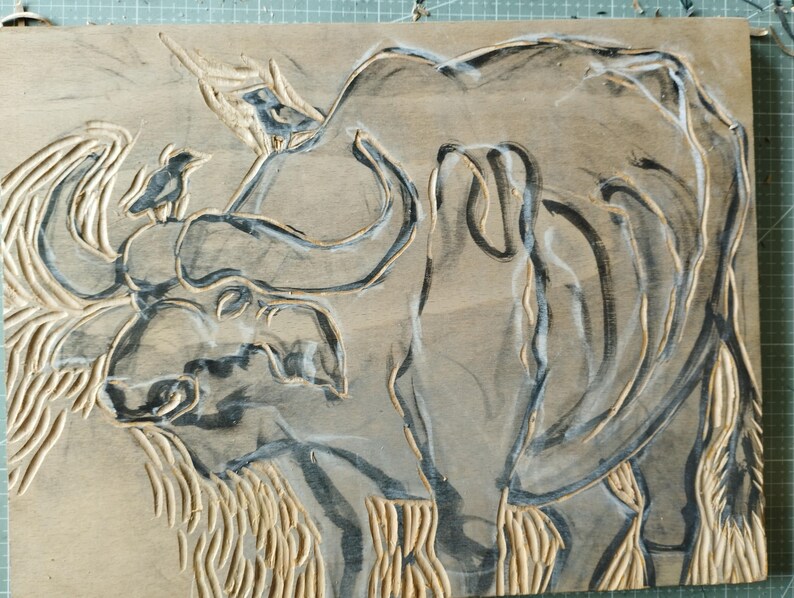 May include: A rectangular wooden block featuring a carved relief of a large buffalo with a small bird perched on its head. The design is highlighted with dark ink within the carved grooves, contrasting against the natural light brown wood surface.