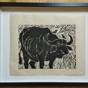 May include: A black and white linocut print of a buffalo on cream-colored paper, framed in a black frame. The artwork features a buffalo with two small birds on its back, surrounded by stylized grass patterns. The text "Buffalo" is printed below.