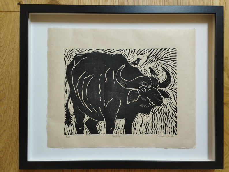 May include: A black and white linocut print of a buffalo on cream-colored paper, framed in a black frame. The artwork features a buffalo with two small birds on its back, surrounded by stylized grass patterns. The text "Buffalo" is printed below.