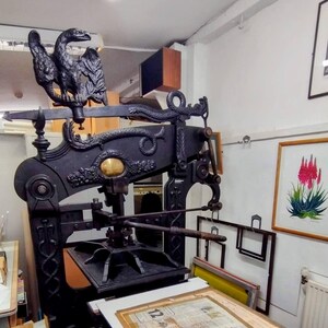 May include: A large, black, cast-iron antique printing press featuring an ornate eagle sculpture at the top and a serpent motif along the horizontal beam. The machine includes a central gold-colored circular emblem and a flat bed with a printed document.