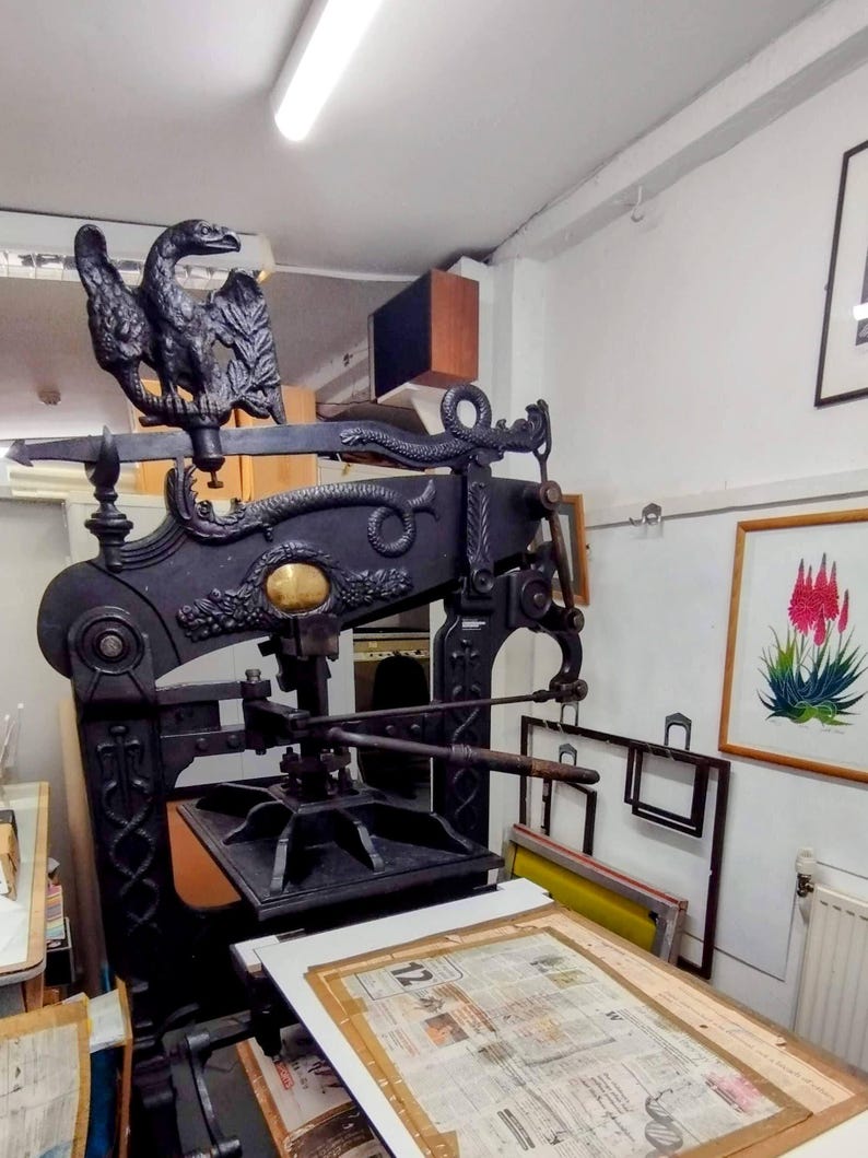 May include: A large, black, cast-iron antique printing press featuring an ornate eagle sculpture at the top and a serpent motif along the horizontal beam. The machine includes a central gold-colored circular emblem and a flat bed with a printed document.