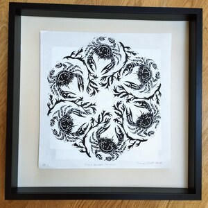 May include: A black and white linocut print featuring a circular mandala pattern of six crabs intertwined with seaweed. The artwork is presented in a square black frame with a white mount. The text "Crab & Seaweed Mandala" is printed at the bottom.