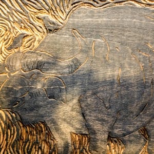 May include: A woodblock print featuring a carved image of a large buffalo with curved horns. The artwork displays deep grooves and textured lines throughout the surface, creating a high-contrast visual effect with dark blue and natural wood tones.