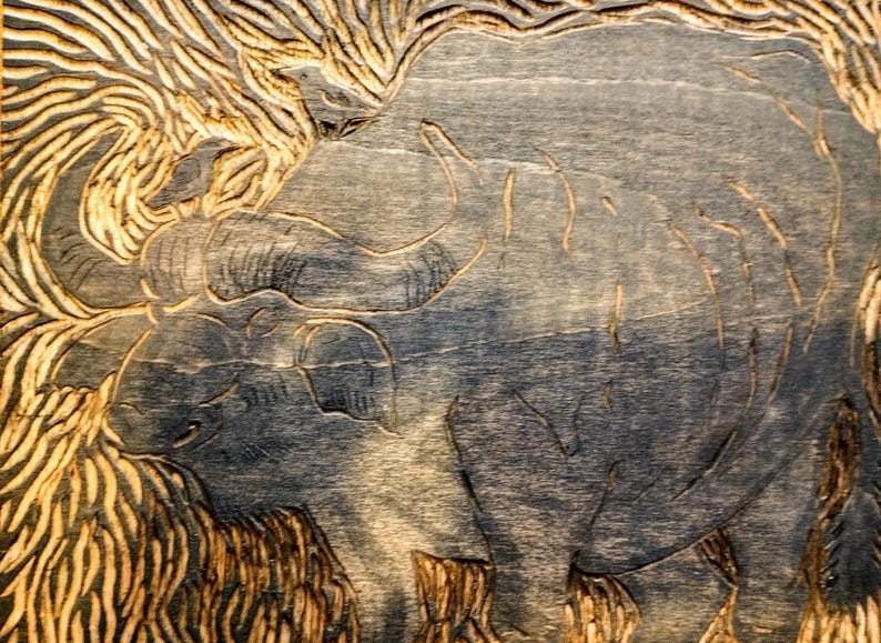 May include: A woodblock print featuring a carved image of a large buffalo with curved horns. The artwork displays deep grooves and textured lines throughout the surface, creating a high-contrast visual effect with dark blue and natural wood tones.