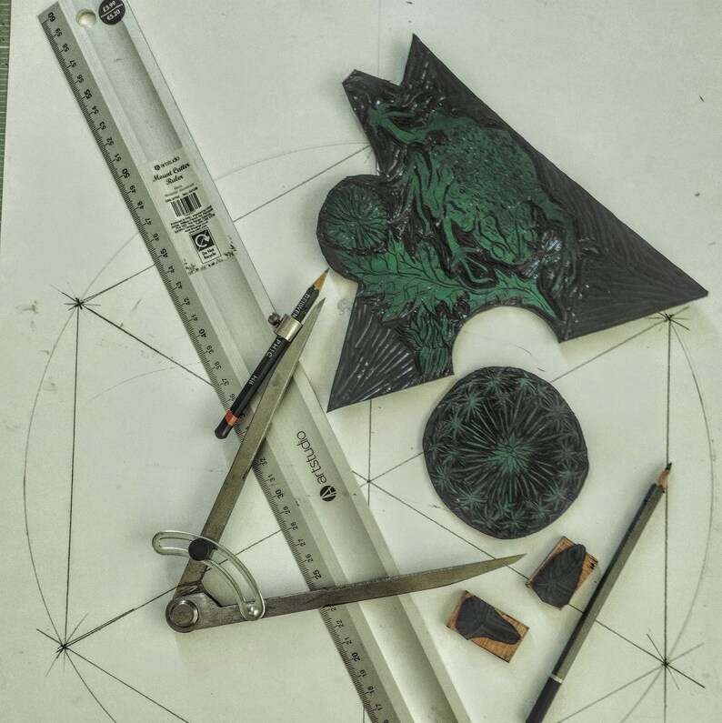 Pu&ograve; includere: Un set di blocchi per linoleografia con un design triangolare di rana e un motivo circolare di tarassaco, accompagnati da un compasso in metallo, un righello da 30 cm e due matite in grafite su una superficie bianca con schizzi geometrici.