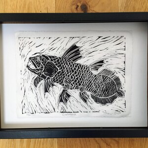 Black and white relief print of a coelacanth fish swimming through textured water marks. 
Printed on hand torn deckle edged Wenzhou paper. Features detailed scales, lobed fins, and hand-penciled signature and edition details along the bottom margin. Displayed in a black frame