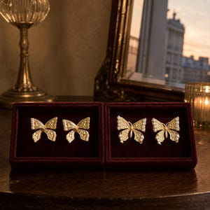 May include: Two pairs of gold-toned butterfly earrings displayed in dark red velvet boxes. The first pair features a textured, solid gold-colored butterfly design, while the second pair features a similar butterfly shape accented with small white stones.