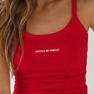 Könnte beinhalten: Eine anthrazitfarbene Baseballkappe und ein leuchtend rotes Ripp-Top mit dem weißen Schriftzug "MATCHA MY ENERGY" auf der Brust, kombiniert mit einer hellblauen Jeans. Das Top hat einen Rundhalsausschnitt und breite Schulterträger.