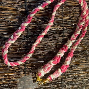 Op de afbeelding: Een gevlochten koord in roze, lichtroze en witte tinten, voorzien van een doorzichtig rechthoekig plastic lipje voor telefoonbevestiging aan de onderzijde, vastgezet met twee goudkleurige metalen karabijnsluitingen die het koord verbinden.