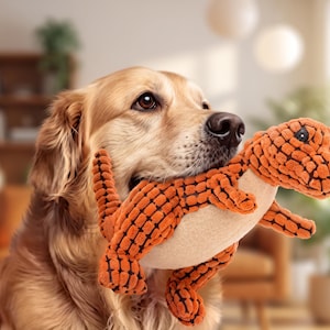 Puede incluir: Un perro de color dorado que sostiene en su boca un juguete de peluche con forma de dinosaurio naranja y crema. El juguete presenta un patrón texturizado de rejilla en su exterior naranja, una barriga lisa de color crema y un ojo negro.