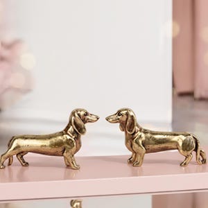 May include: Two metallic gold-toned dachshund figurines standing in profile, facing each other. Each dog sculpture features a smooth, polished finish with subtle dark shading in the crevices to highlight the anatomical details of the ears and body.