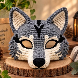 May include: A handcrafted crochet wolf mask featuring a grey and cream colour palette with black accents on the nose and forehead markings. The mask has pointed ears, almond-shaped eye openings, and a textured yarn construction throughout.