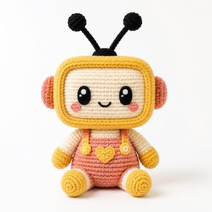 May include: A crocheted plush toy shaped like a television set with a yellow frame, cream-colored screen, and black antennae. The figure wears pink overalls with a yellow heart detail, pink side knobs, and has large black eyes with white highlights.